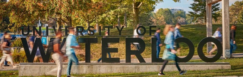 ‘UNIVERSITY OF WATERLOO’라는 대형 입간판 앞을 학생들이 분주하게 지나가고 있다. 가을 햇살이 비추는 캠퍼스 배경에는 초록 나무들과 잔디, 그리고 멀리 보이는 건물들이 조화를 이루고 있다.