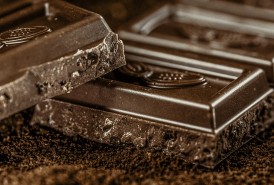 두껍고 네모난 모양의 다크초콜릿을 클로즈한 사진으로 바닥에는 초콜릿가루가 가득하다.