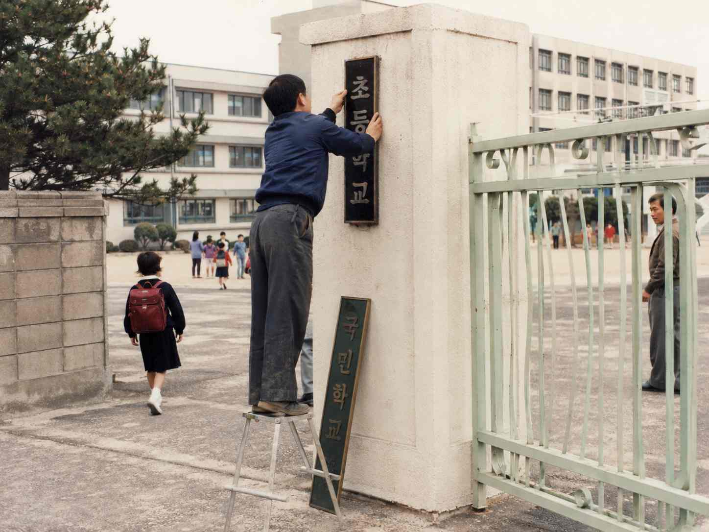 오래된 학교 정문에서 기존 명패를 떼어내고 새로운 명패를 준비하는 모습을 재현한 장면