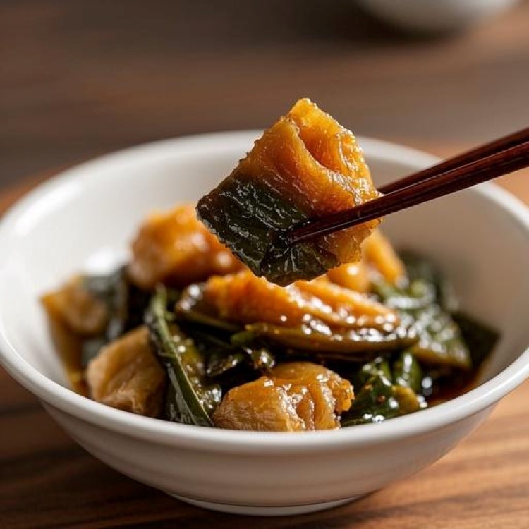 윤기 나는 갈색 머위대볶음이 하얀 그릇에 담겨있는 모습, 들깨가루의 고소한 느낌이 묻어나는 색감, 젓가락으로 집어 올린 부드러운 머위대의 질감이 보이는 한식 요리 사진, 자연광 아래서 촬영