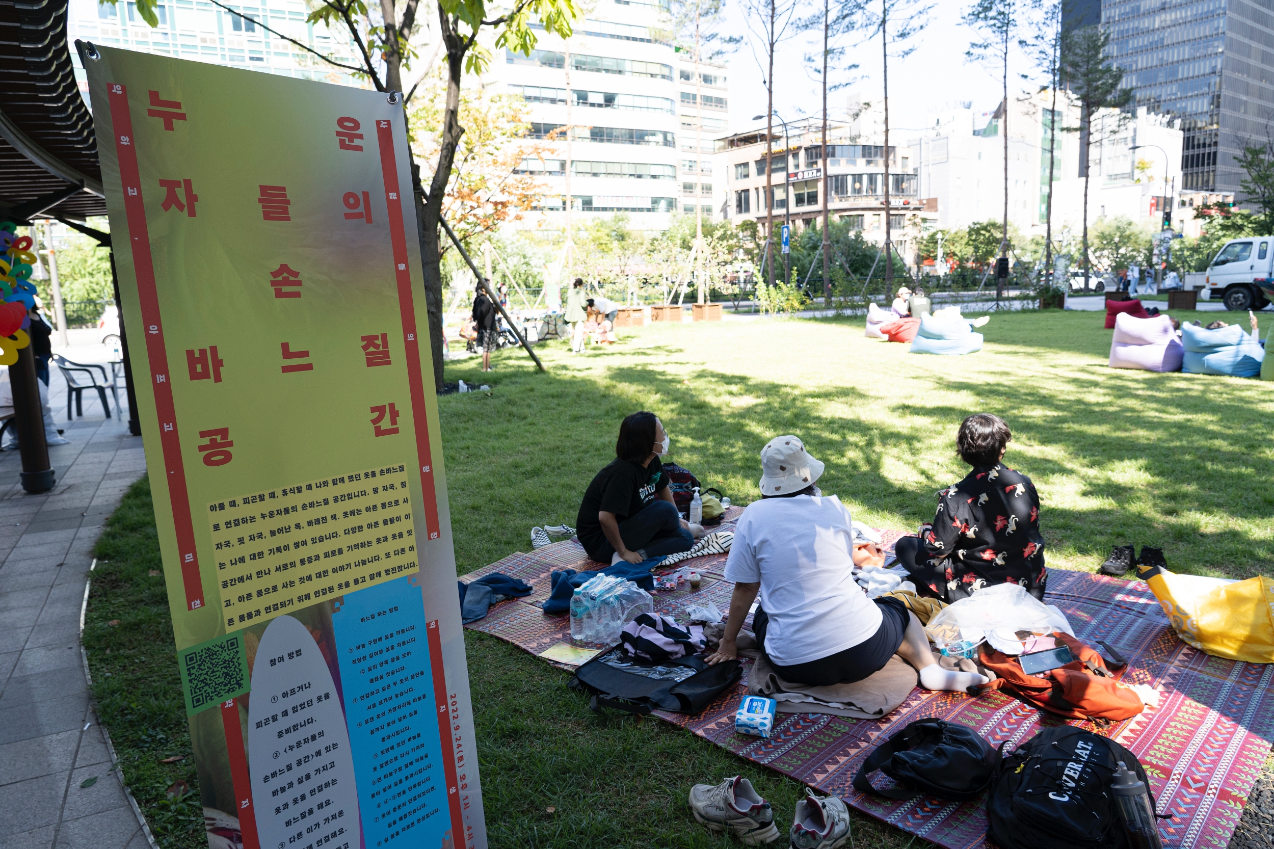 <누운 자들의 손바느질 공간> 배너 옆으로 시민들이 잔디밭에 앉아 손바느질을 하고 있다.