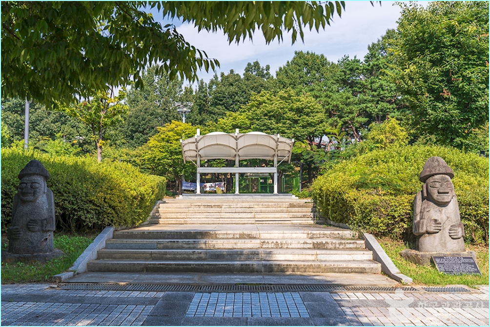 계단 양 옆으로 돌하르방이 서 있고 중앙에 하얀 지붕의 쉼터가 보이는 발산공원 풍경.