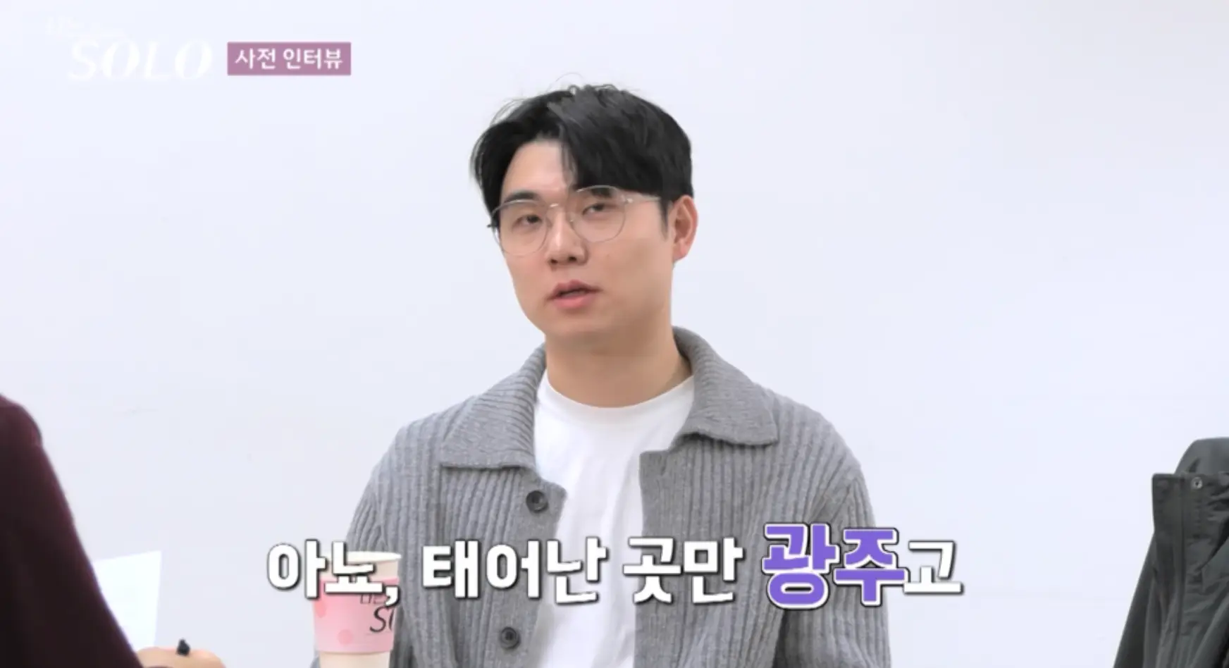 나는 솔로-회색니트 안에는 흰색 티를 입고 커피를 든채 미소지으며 말하는 상철 아래 흰색과 보라색이 섞인글시 아뇨. 태어난 곳만 광주고