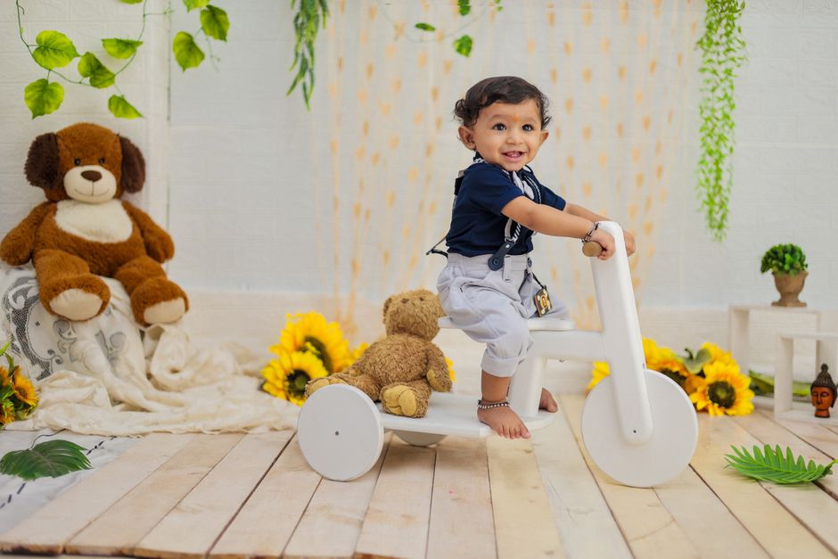 A joyful toddler rides a white tricycle surrounded by plush toys and greenery decor indoors.