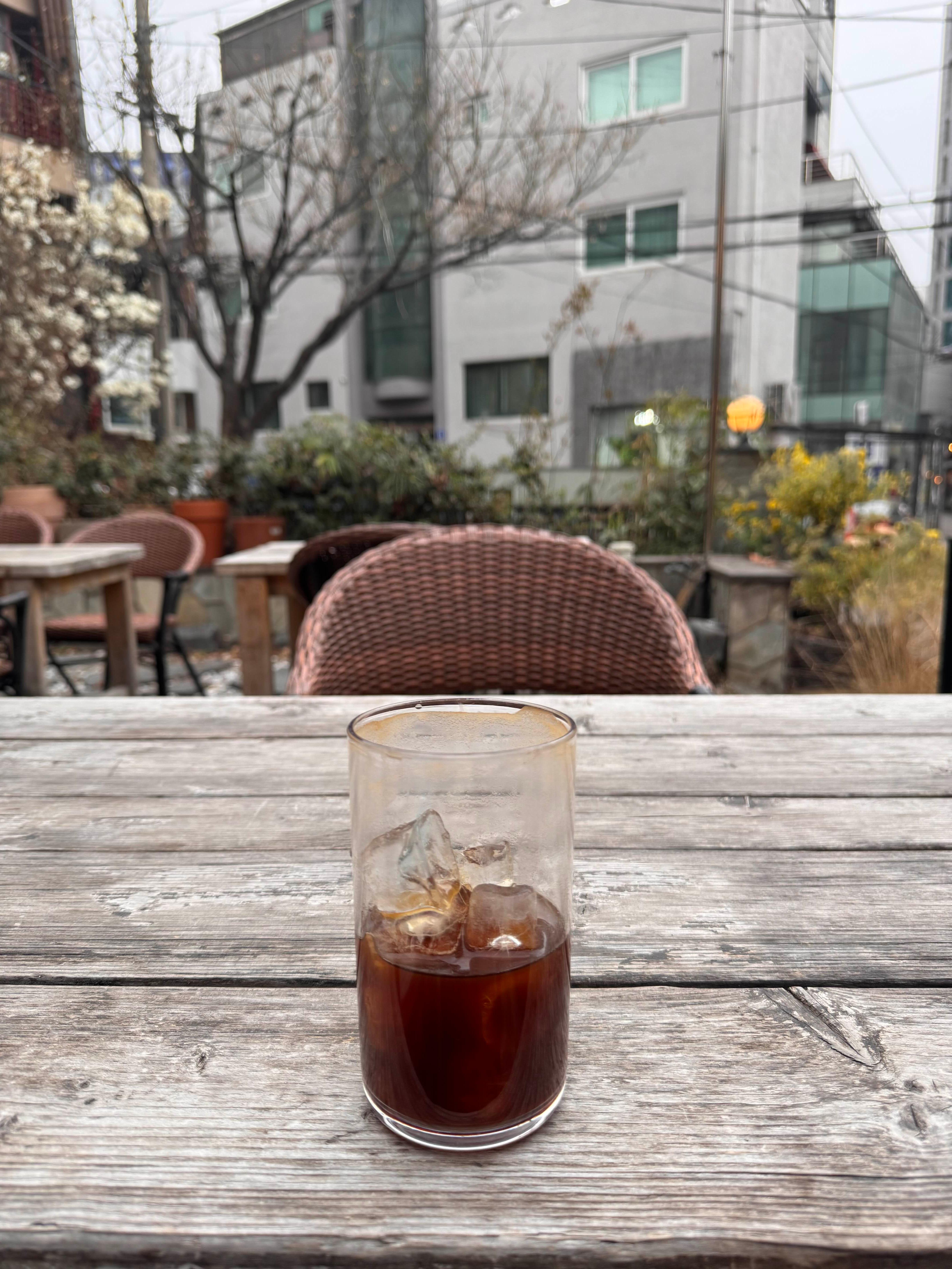 야외 테라스 나무 테이블 위에 놓인 투명한 유리컵과 뒤로 보이는 고즈넉한 도시 건물의 풍경
