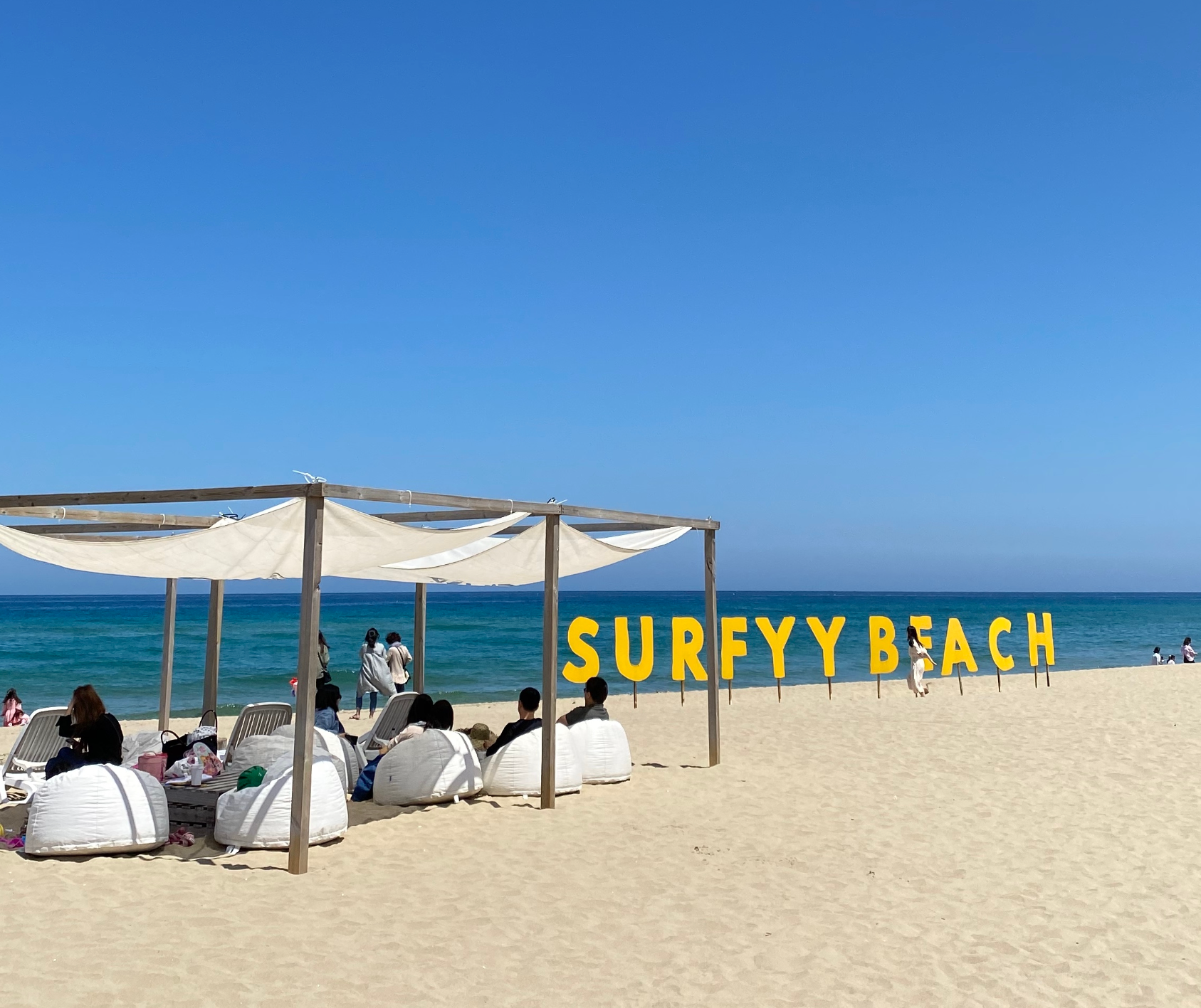 강원 양양 서피비치에서 해변 파라솔 아래 앉아 바다를 감상하는 사람들과 노란색 SURFYY BEACH 사인
