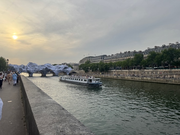A scenic view of the Seine river in Paris with a cruise boat and the wrapped Pont Neuf bridge under a cloudy sunset sky ❘ Une vue panoramique de la Seine &agrave; Paris avec un bateau de croisi&egrave;re et le pont du Pont-Neuf envelopp&eacute; sous un ciel de coucher de soleil nuageux⁠