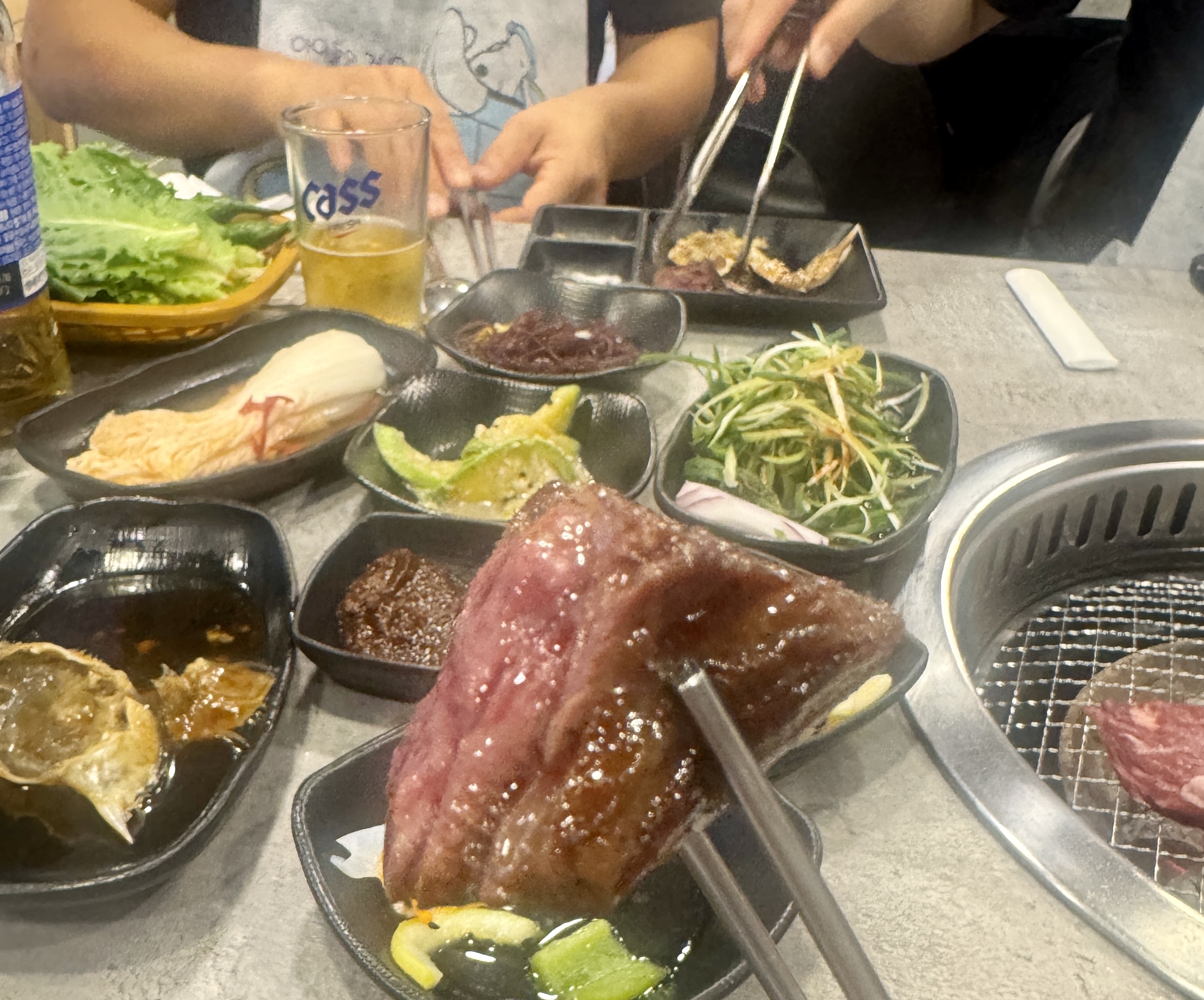 삼거리 명품한우에서 숯불에 구운 한우 한 점을 젓가락으로 들어 올린 모습. 육즙이 촉촉하게 배어 있으며 배경엔 밑반찬과 맥주가 놓여 있다.