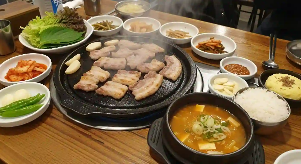 삼겹살이 맛있게 구워지고 있고, 쌈과, 된장찌개, 김치, 마늘이 있는 테이블 모습