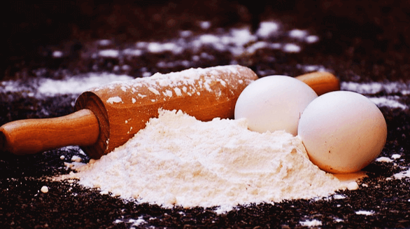 검정 탁자에 수제비 재료인 하얀 밀가루, 하얀 달걀 2개, 나무 밀대가 있다.