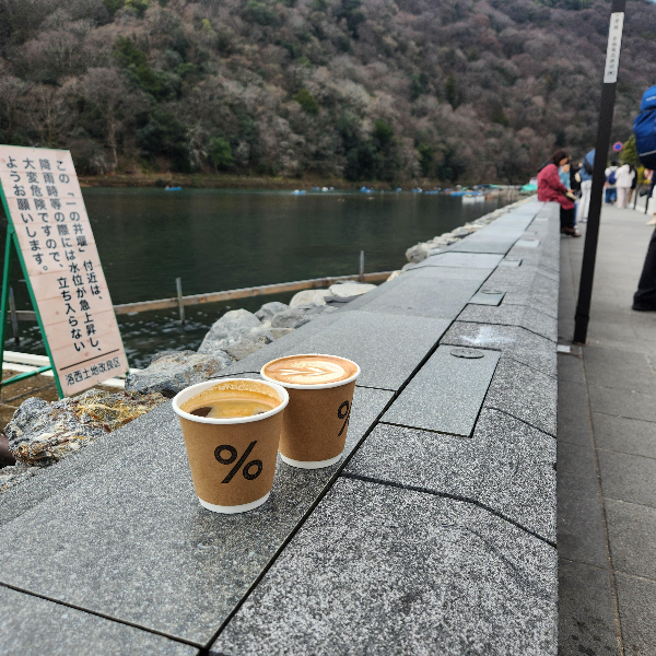 강가 제방 위에 커피 두 잔이 올려져 있다. 컵에는 %로고가 프린트 되어 있다.