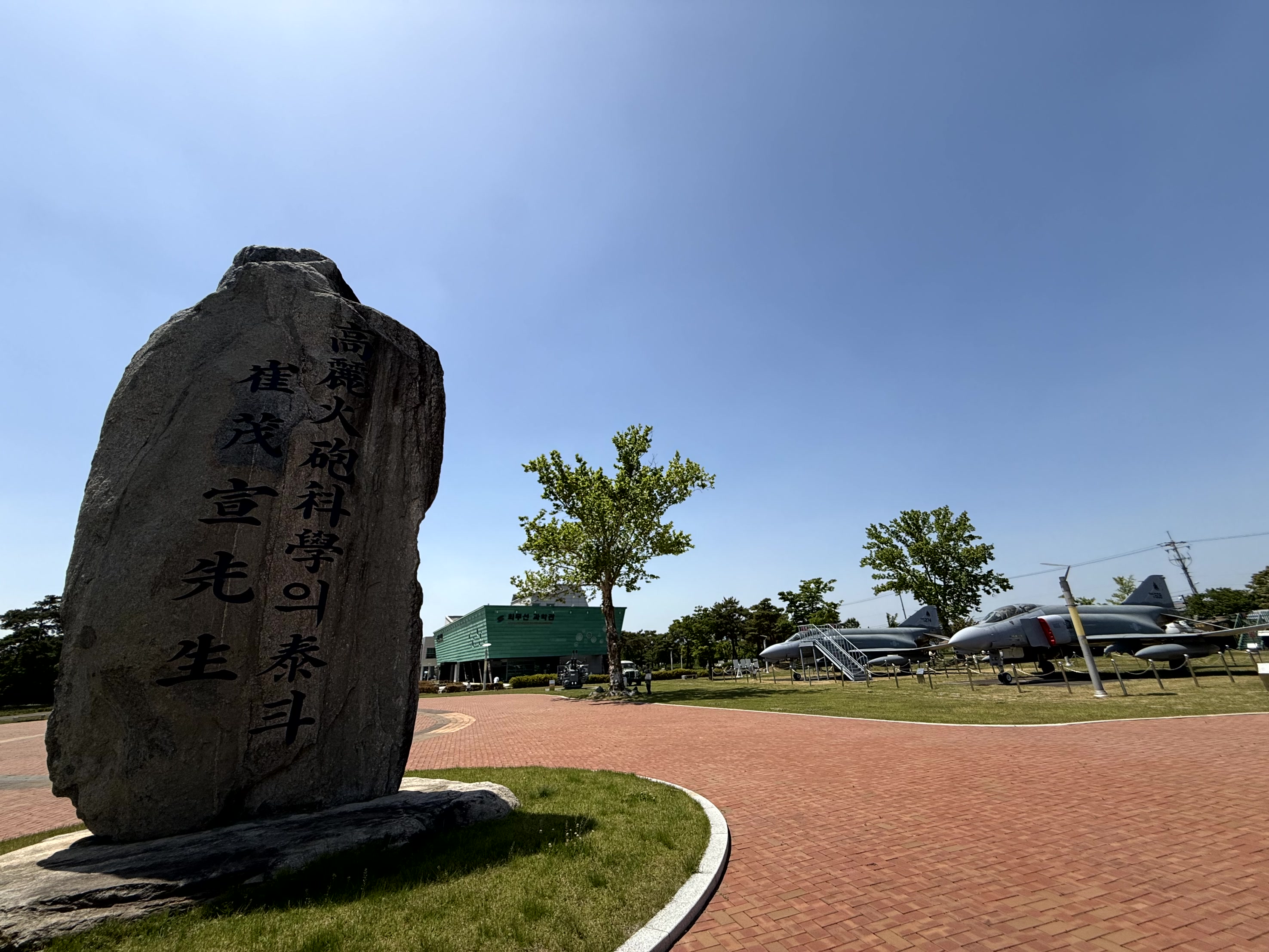 영천 최무선과학관 야외 전시장. 커다란 비석이 돋보이며, 붉은 벽돌길과 푸른 잔디밭이 어우러져 있습니다. 저 멀리 보이는 녹색 건물은 과학관 본관이며, 전투기 전시물도 보입니다.