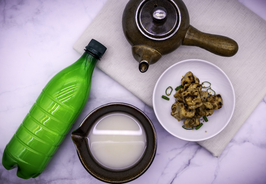 흰색 바닥 위에 초록색 먹걸리 병을 눕혀 놓고, 그 옆에는 막걸리는 담은 고동색 잔과 고동색 술주전자, 그리고 흰색 그릇에 두부조림을 담아두고 찍은 사진