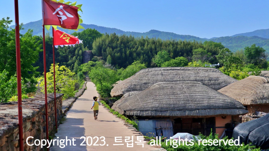 파란하늘과 빨간 깃발에 초가지붕이 보이고 성벽에서 달려가는 노란옷 입은 아이의 뒷모습