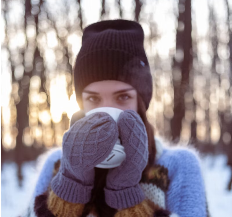 눈 덮인 숲에서 니트 장갑을 낀 여성이 따뜻한 컵을 감싸 쥐고 있는 모습