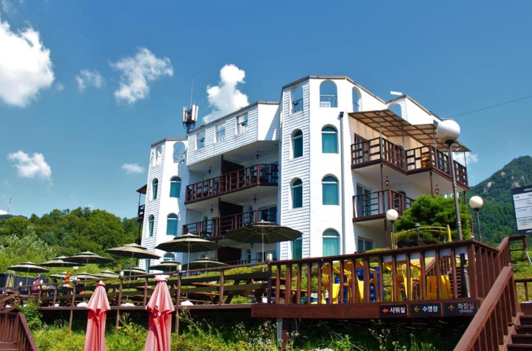 경기도 가평군에 위치한 트래블리 호텔의 외관. 파란 하늘 아래 하얀 외벽과 아치형 창문이 인상적인 건물로, 데크 위 파라솔과 수영장 안내 표지판이 눈에 띈다.