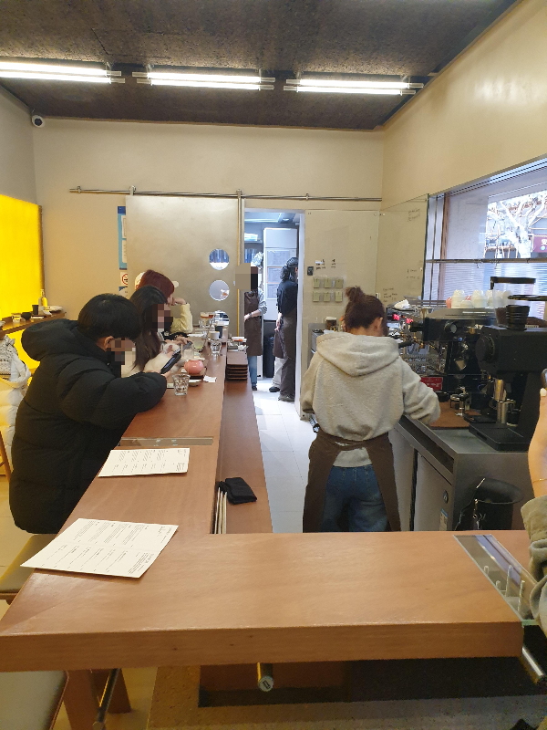 목재로 된 ㄱ자 바 테이블에 고객들이 앉아 있고 바리스타는 업무 중이다.