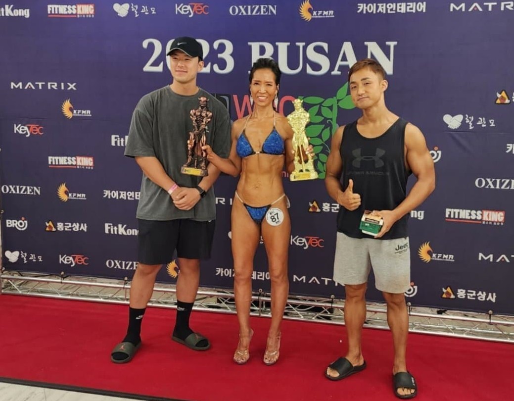 시니어 피트니스에 참가한 박향자씨가 수영복 차림으로 트로피를 양손에 들고 붉은 카펫 위에서 매니저로 보이는 젊은 두 남성과 함께 포즈를 취하고 있는 이미지