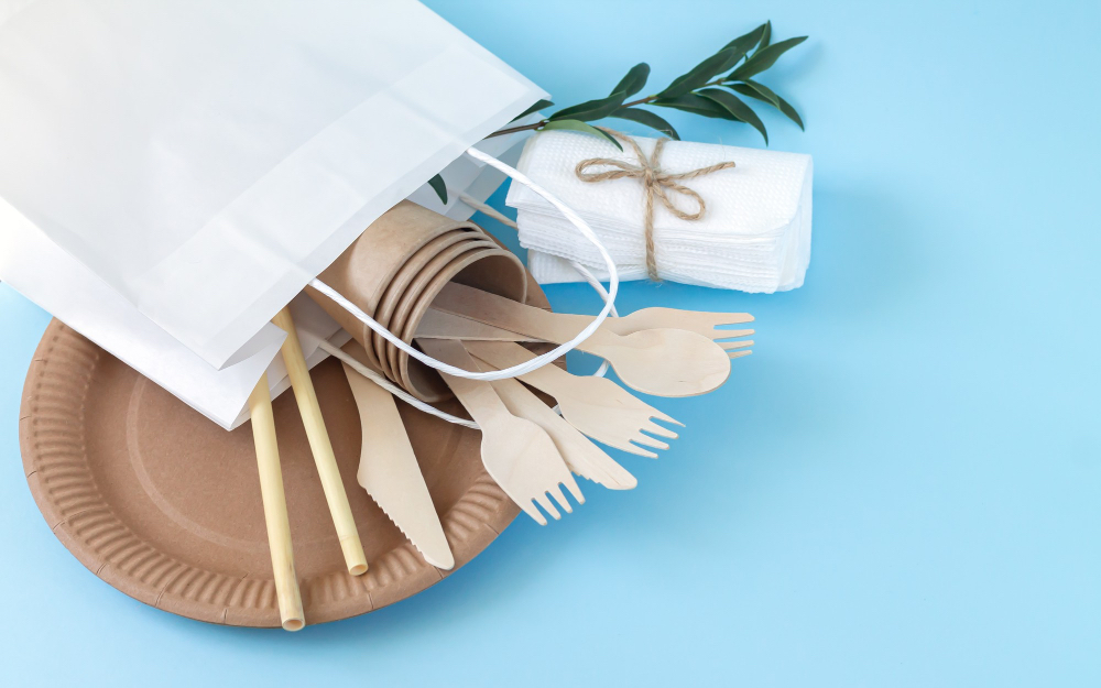 하늘색 배경 위에 친환경 일회용품이 담긴 흰색 종이 쇼핑백이 있습니다. 종이로 만든 접시, 나무로 만든 포크, 나이프, 스푼, 빨대 등의 친환경 일회용 식기구들과 자연 소재로 만든 냅킨이 보입니다. 일회용품 사용을 줄이거나 친환경 소재로 대체하는 방법을 보여주는 이미지입니다.