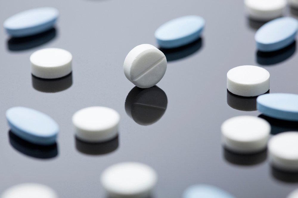 이 사진은 검은색 반사되는 표면 위에 흩어진 여러 개의 알약을 보여줍니다. 알약들은 흰색과 파란색으로 구성되어 있으며, 일부는 둥글고 일부는 타원형입니다. 중앙에는 홈이 파인 흰색 알약이 세워져 있어 강조된 모습입니다.