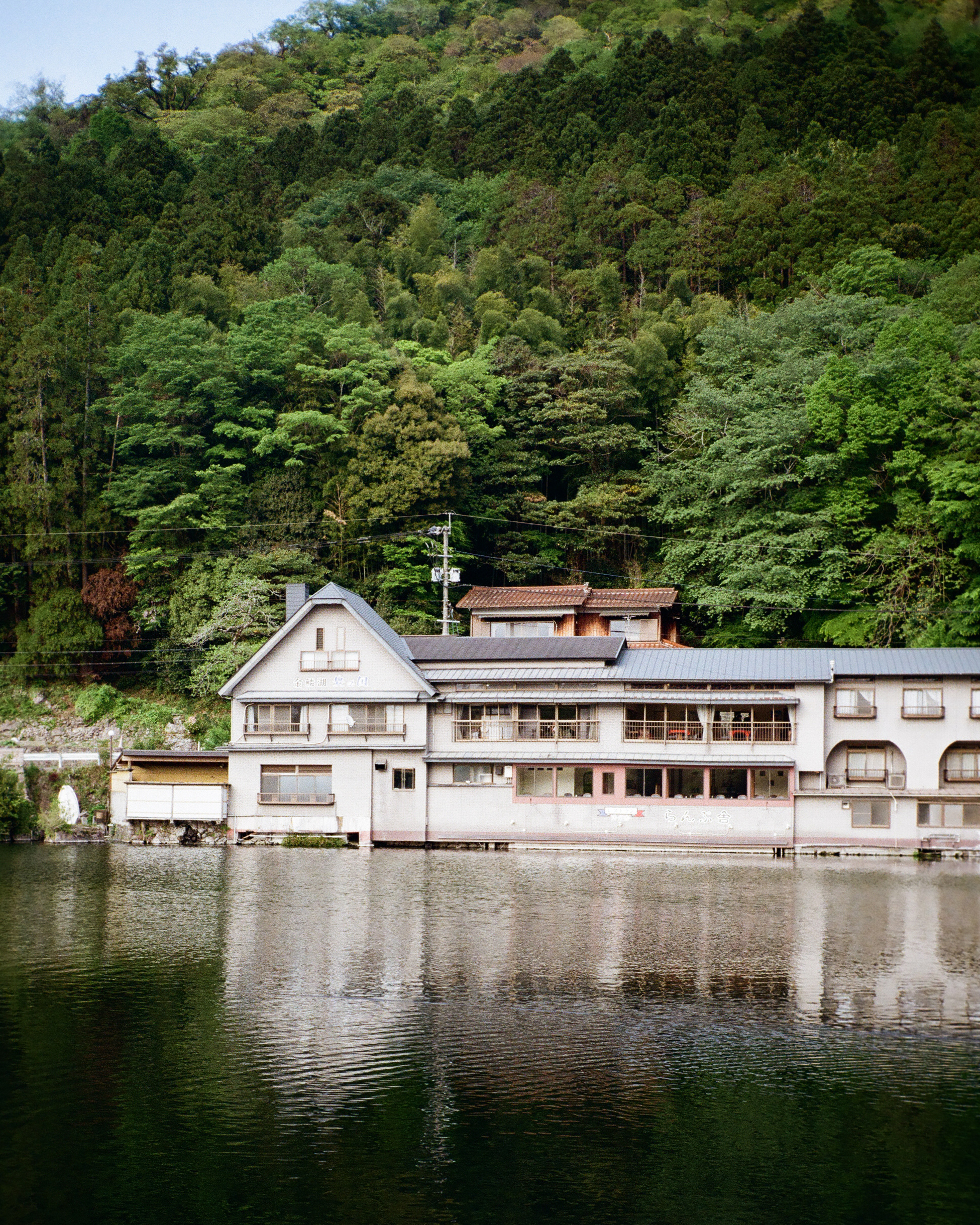 잔잔한 호숫가에 위치한 일본풍 건물들과 그 뒤를 감싸고 있는 울창한 초록빛 산의 풍경. 건물들이 수면에 맑게 투영되고 있으며, 전체적으로 포트라 400 필름 특유의 따뜻하고 부드러운 녹색조와 차분한 베이지색 톤이 조화를 이룸. 필름의 질감이 살아있는 선명하면서도 편안한 풍경 사진이다.