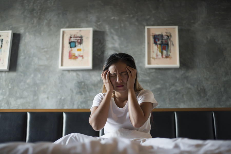 흰색 반팔 셔츠를 입은 머리가 긴 서양 여성이 침대에 앉아서 두통을 호소하며 양쪽 손으로 머리를 감싸고 있는 것을 찍은 사진