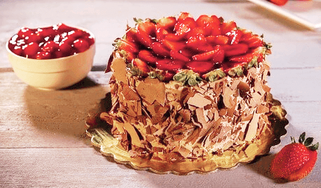 나무 탁자 위에 동그란 초콜릿 생일 케이크가 있다. 맨 위에는 빨간 딸기들로 장식됐고 옆에 그릇에는 체리가 한가득 있다.