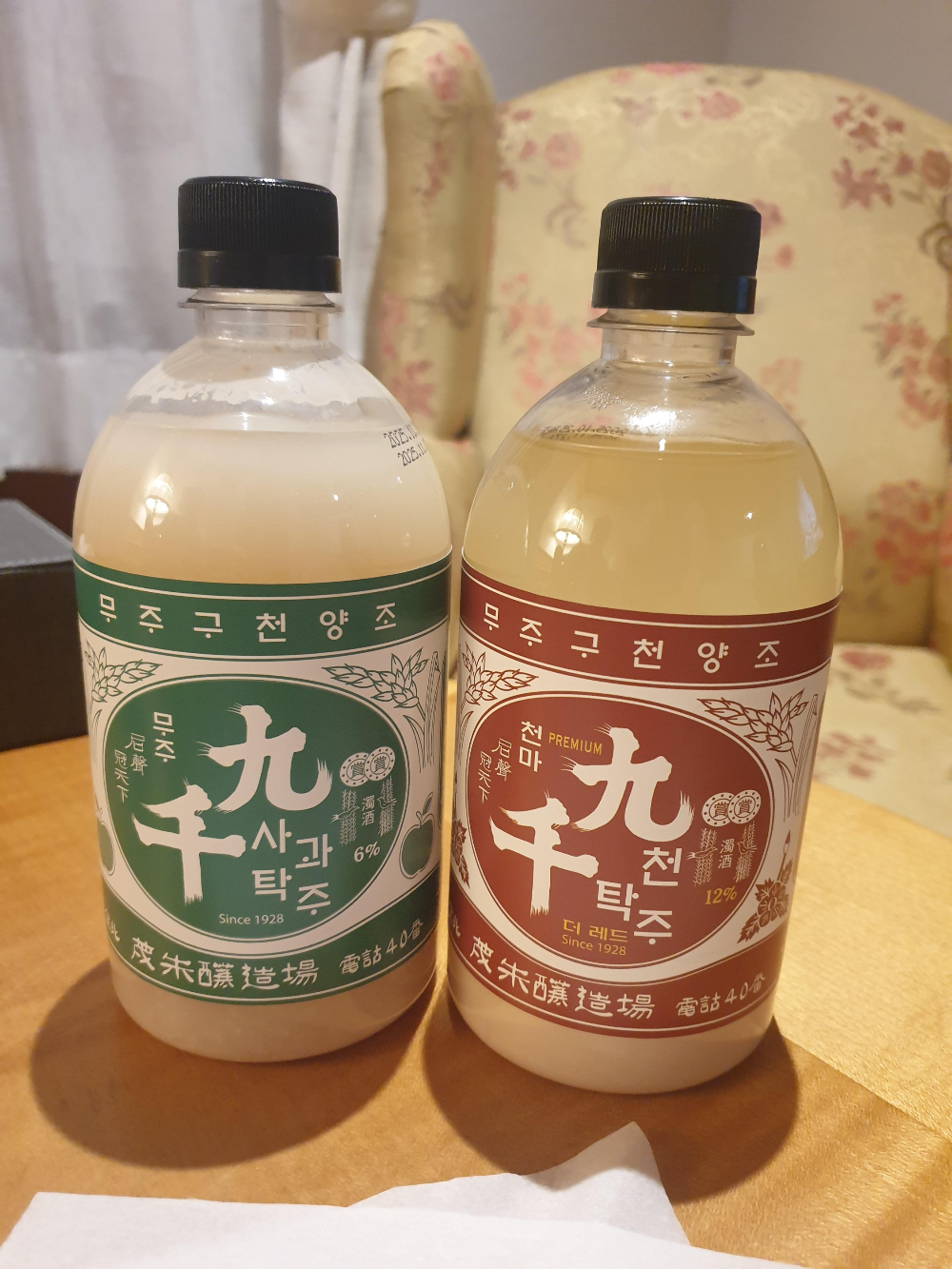 호텔 둥근 목재 테이블 위에 탁주 2병이 놓여 있다. 하나는 사과탁주로 초록 라벨 오른쪽은 붉은 라벨의 천탁주레드다.