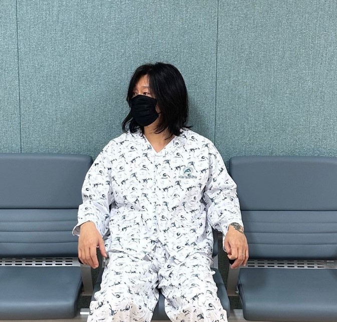 윤도현 씨가 의자에 앉아 오른쪽을 쳐다보고 있다. 검정 마스크를 쓰고 흰 색 환자복을 입고 있음