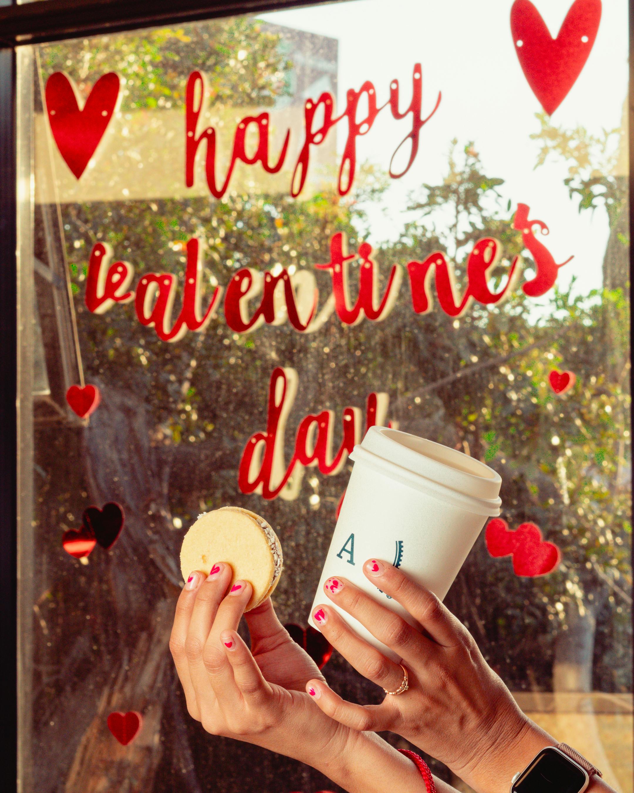 발렌타인 데이 글씨가 적힌 창문과 그 앞에 커피와 마카롱을 들고있는 손 모습