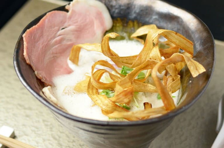 하얀색 거품이 그릇안에 있고 그 위로 노란 얇은 튀김과 차슈가 올라가 있다