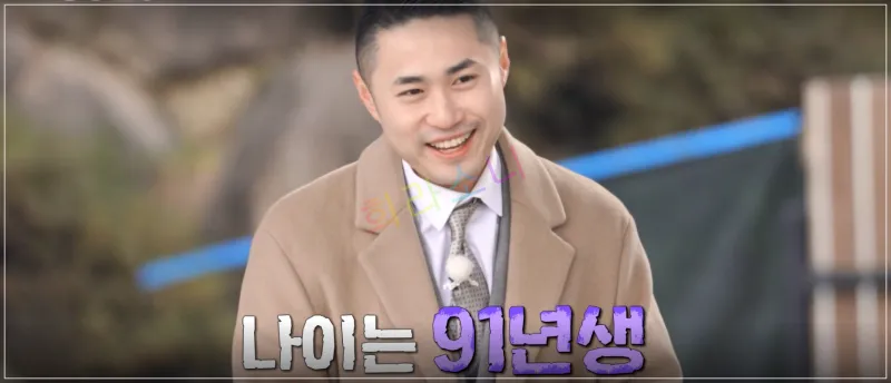 영수가 환하게 웃으며 서 있는 모습이다. 배경에는 자연 풍경이 보이며&amp;#44; 그는 베이지색 코트에 넥타이를 맨 정장 차림이다. 자막: 