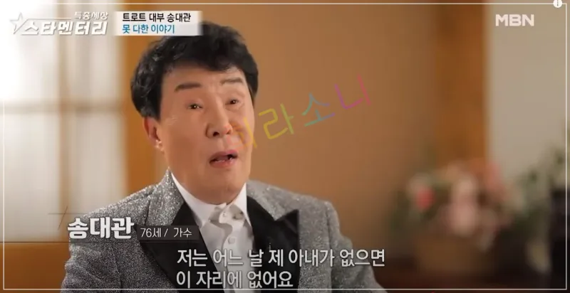 흰 머리와 검은 머리칼이 섞인 송대관이 반짝이는 은색 정장을 입고 인터뷰를 하는 모습. 화면에는 &amp;quot;저는 어느 날 제 아내가 없으면 이 자리에 없어요&amp;quot;라는 자막이 있다. 배경은 따뜻한 조명 아래 실내 공간이다.
