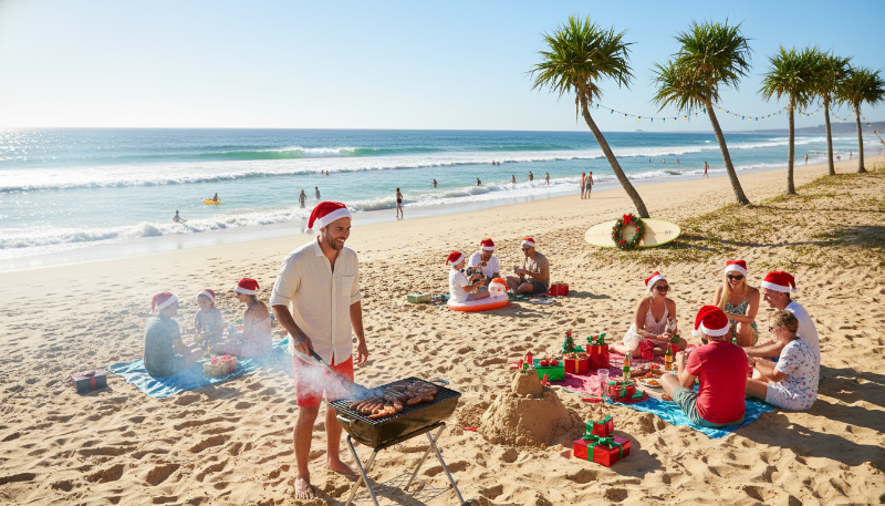 햇살 가득한 호주 해변에서 크리스마스를 즐기는 사람들. 모래사장에서 바비큐를 하고 물놀이를 하며, 산타 모자를 쓴 사람이 그릴 앞에서 요리하고 있습니다.