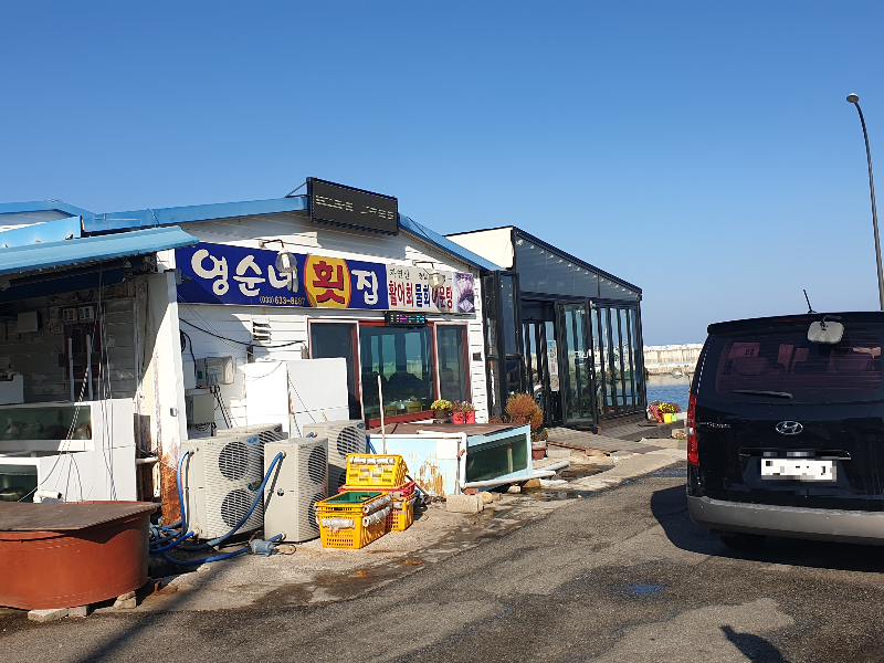 파란 하늘 아래 두 개의 건물이 이어져 있다. 앞 쪽 건물에 간판이 있다. 그 앞을 차량 한 대가 지나간다.