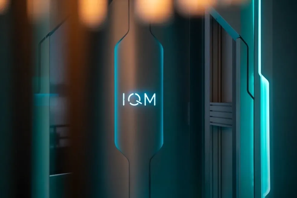 IQM 양자컴퓨터 외부 하우징. 청록색 LED 조명 아래 IQM 로고가 새겨진 중앙 타원형 패널과 정밀 냉각 구조물이 보이는 양자컴퓨팅 하드웨어 시스템이다.