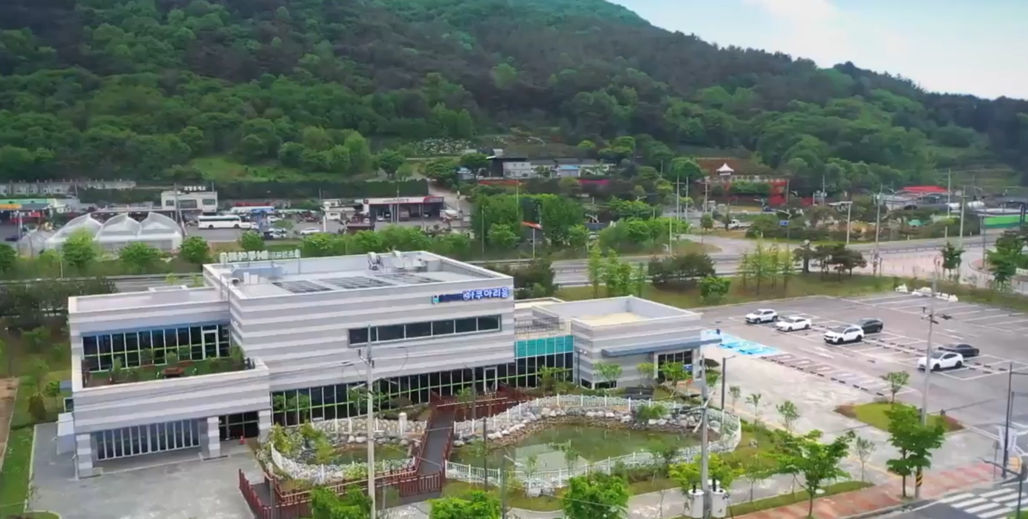 충북 괴산 충북아쿠아리움 외관 항공 뷰. 숲으로 둘러싸인 현대적인 건물과 앞쪽 연못, 넓은 주차장이 함께 보이는 모습