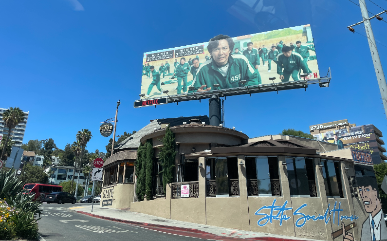 낮은 건물 지붕 위에 큰 광고판이 세워져 있다. 광고판에는 오징어 게임의 주인공인 이정재 배우와 다른 배우들이 모두 같은 초록색 저지를 입고 있는 모습이 나타나 있고, 아래쪽으로 넷플릭스라고 쓰여져 있다.