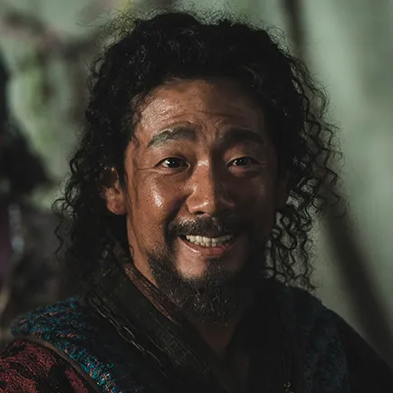 꼽슬머리를 하고 있는 30대 남성이 짙은 빨강과 청색 옷을 입고 서 있는 아라문의 검 쇼르자긴 캐릭터