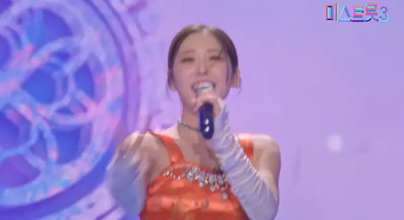 미스트롯3- 파란배경 앞 주황 나시 티 왼손으로 마이크를 들고 머리를 뒤로 하나로 묶은채 노래하는 윤서령