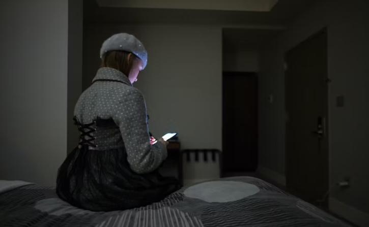 어두운 방 안 침대에 앉아 스마트폰 화면을 바라보는 여성의 뒷모습