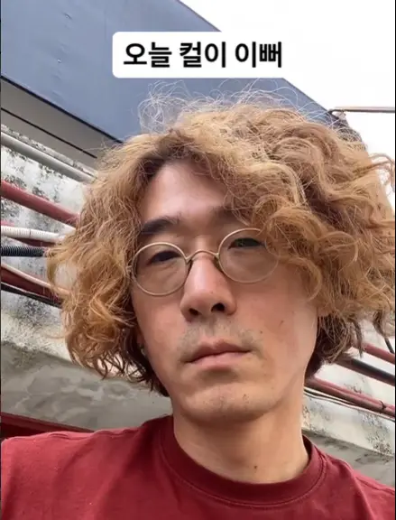 싱어게인3- 야외 붉은 티셔츠 노란색 곱슬거리는 머리에 무심한 표정으로 머리맡 흰바탕검은글씨 오늘은 컬이 이뻐