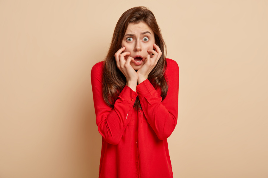 빨간 셔츠를 입은 긴머리의 젊은 서양 여성이 양쪽 손을 턱부분에 대고 깜짝 놀라고 있는 것을 찍은 사진