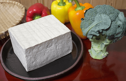 갈색 도자기그릇에 커다랗고 하얀 두부 한 모가 있고, 오른쪽에는 브로콜리와 빨갛고 노란 파프리카들이 있다.