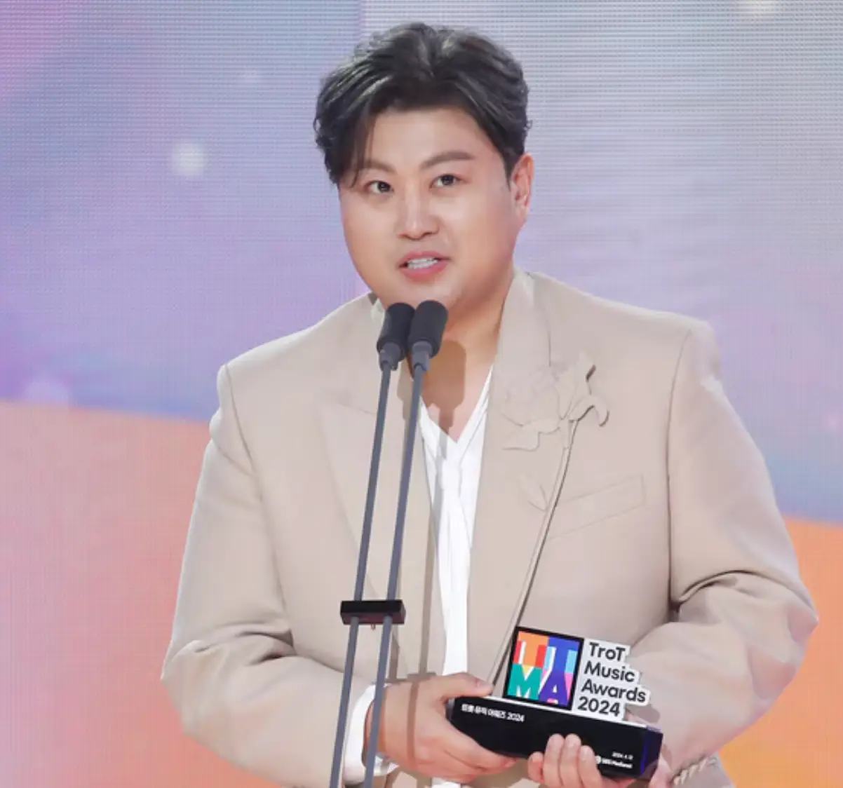 아이보리색 쟈켓 트로피를 들고 스탠딩 마이크 앞에서 미소지으며 말하고 있는 김호중