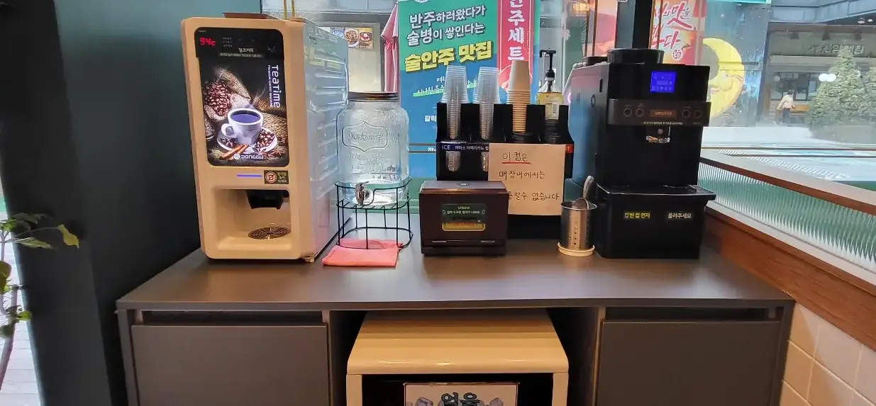보배반점 -
나무 테이블 위 흰색 믹스커피 자판기 옆 물이 가득 담긴 투명 생수병 옆 종이컵과 플라스틱 컵이 가득 쌓인 컵 홀더 옆 아메리카노 커피 자판기