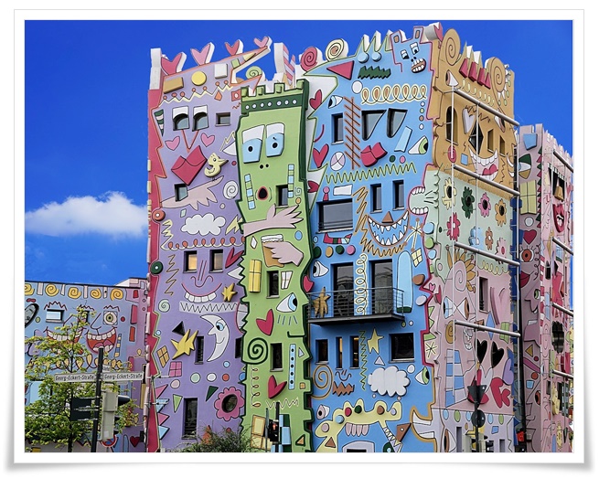 밝은 푸른 하늘을 배경으로 파스텔 퍼플, 그린, 블루, 핑크 색상의 만화 얼굴과 하트, 별, 달등을 소재로 하는 기발한 팝 아트 디자인으로 장식된 건물 전경