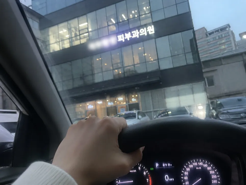 차 안에서 운전대를 잡고 전면 유리창 너머로 불이 켜진 피부과 의원 건물을 바라보고 있는 내돈내산 피부과 방문 인증 사진