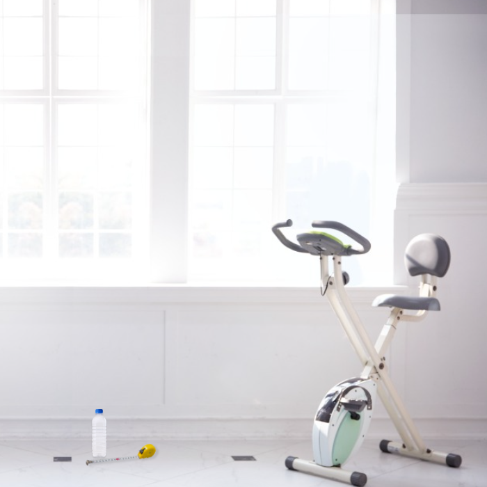 햇빛이 화사하게 드는 창가에 실내 자전거가 오른쪽에 놓여 있고 왼쪽에 생수와 자가 놓여 있는 이미지