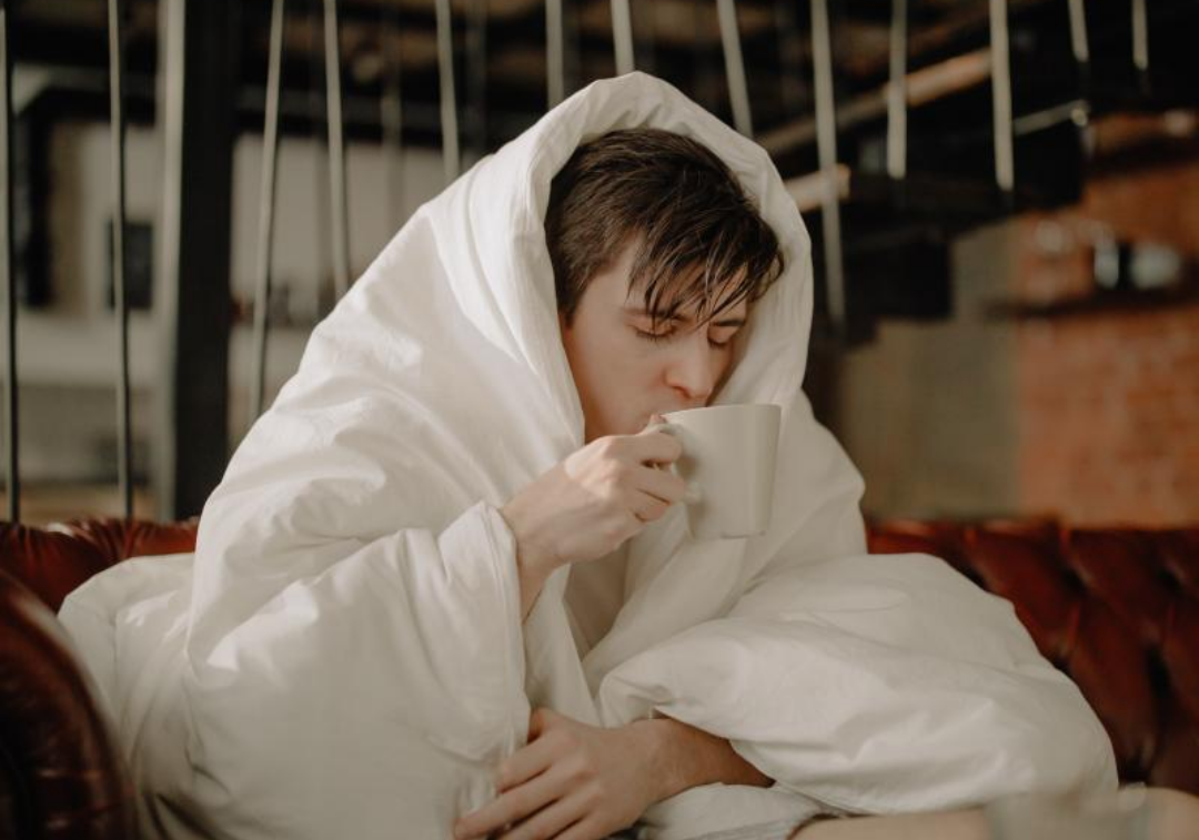 몸살에 걸린 남자가 이불을 쓰고 차를 마시고 있다.